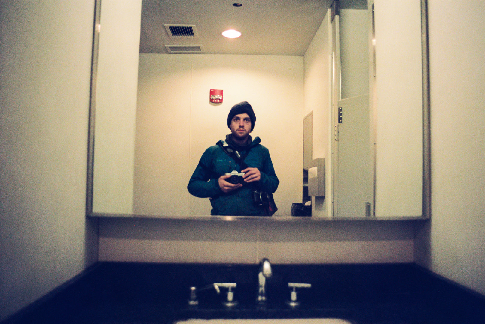 A man wearing a blue hooded rain jacket and a black beanie stands in a bathroom, captured in a mirror reflection above a dark sink. The photo has a cool, slightly filtered tone.
